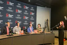 Thumbnail for Panel of four people with headphones behind microphones facing a standing woman speaking into a microphone in a recording studio