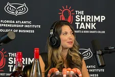 Thumbnail for Woman with headphones sits at podcast table with shrimp candy bowl, drinks, and cap, with overlaid text reading The Shrimp Tank.
