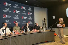 Thumbnail for Panel of four men with headsets at a desk facing a woman standing with papers in a podcast studio with branded backdrop.
