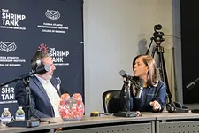 Thumbnail for Two people recording a podcast episode at a table with microphones and a backdrop for The Shrimp Tank show.