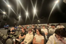 Thumbnail for Group of young adults seated attentively in a dimly lit theater or lecture room with black walls and bright ceiling lights.
