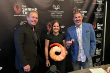 Thumbnail for Three people posing at a Shrimp Tank event backdrop, one holding a large shrimp plush, with drinks and microphones nearby.