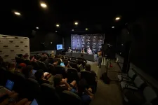 Thumbnail for Audience attentively watching a panel discussion in a dark conference room with speakers at a table and media equipment.