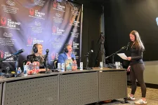 Thumbnail for Radio hosts in headphones recording a podcast with a standing woman speaking into a microphone in a studio.
