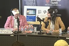 Thumbnail for Two podcast hosts wearing headphones converse at a table with microphones and shrimp decor, Shrimp Tank event backdrop.