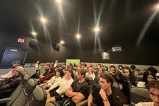 Thumbnail for Audience seated and attentively watching in a small, dark theater with overhead spotlights.