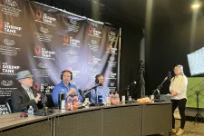 Thumbnail for Podcast recording with three men wearing headphones, microphones, and a woman standing by light and camera setup.