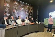 Thumbnail for Panel of five men with headsets seated at a long table during The Shrimp Tank event, woman standing nearby with a phone.