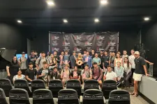 Thumbnail for Large group of young adults posing in a small theater with a Shrimp Tank branded backdrop and seats in foreground.