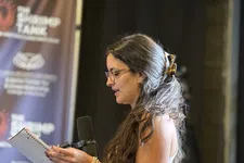 Thumbnail for Young woman with glasses reading from a notebook into a microphone in a recording studio with banners.