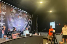 Thumbnail for Podcast recording with four hosts behind microphones and two people standing near a camera and light setup.