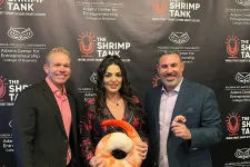Thumbnail for Three people pose smiling in front of a Shrimp Tank branded backdrop, one holding a large shrimp-shaped item.