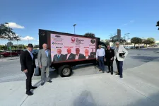 Thumbnail for Six men in business attire stand outdoors beside a large digital screen advertising The Shrimp Tank event.