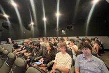 Thumbnail for Group of students attentively seated in a dark lecture hall with black walls and ceiling lights.
