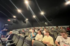 Thumbnail for Group of people seated in a small, dimly lit theater or lecture room with black walls and ceiling lights.