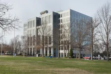 Thumbnail for Modern office building with glass windows behind leafless trees and a green lawn under cloudy sky