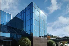 Thumbnail for Modern glass office building reflecting blue sky with white clouds and surrounding greenery.