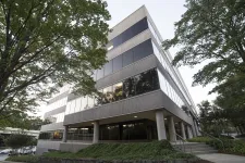 Thumbnail for Modern office building surrounded by green trees with reflective windows and a sidewalk entrance.