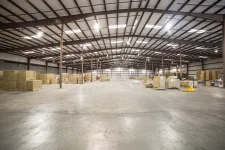 Thumbnail for Spacious warehouse interior with concrete floor, metal beams, and stacks of cardboard boxes under bright lighting.