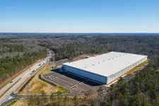 Thumbnail for Aerial view of large white industrial warehouse surrounded by forest and adjacent to a road under clear sky
