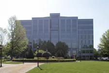 Thumbnail for Modern glass office building with landscaped garden, trees, and lamp posts in the foreground on a sunny day