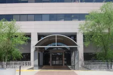 Thumbnail for Modern office building entrance with arched canopy, surrounded by trees and concrete planters on a sunny day