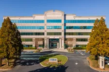 Thumbnail for Modern four-story office building with reflective windows, surrounded by trees under a clear blue sky.