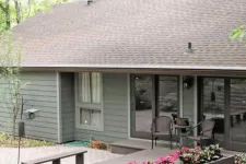 Thumbnail for Cozy gray house with large windows, patio seating, and vibrant pink flowers in outdoor planters under green trees.