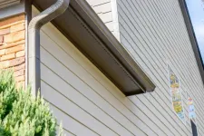 Thumbnail for Close-up of house exterior showing beige vinyl siding, brown gutter system, and part of a brick wall with green shrub.