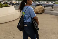 Thumbnail for Woman in blue shirt and jeans standing outdoors with a black backpack and jacket, smiling and looking away.