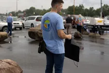 Thumbnail for Young man in blue t-shirt and jeans holding a jacket standing in wet parking lot near large rocks and cars.