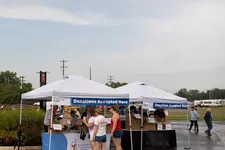 Thumbnail for People donating items at outdoor tents with signs reading Donations Accepted Here on a cloudy day.