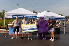 Thumbnail for Group of people at donation tents holding a sign promoting Telhio Day at the Fair 2026 with a purple mascot character.