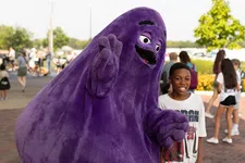 Thumbnail for A boy posing and smiling next to a large, friendly purple mascot in an outdoor public area with people in the background.