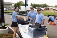 Thumbnail for Three people in blue shirts packing black drawstring bags outdoors at an event with cars and tents in the background