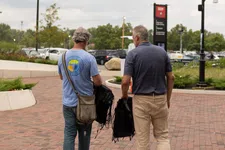 Thumbnail for Two men walking on a brick pathway carrying folded black chairs on a cloudy day in a park-like area.