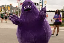 Thumbnail for Person in a large purple mascot costume posing at an outdoor fair with colorful cable cars and people in the background