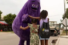Thumbnail for Two children interacting with a person in a large purple mascot costume on a street during daytime.