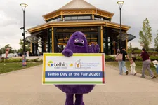 Thumbnail for Purple mascot holding a sign promoting Telhio Day at the Fair 2026 outside a pavilion with people walking nearby.