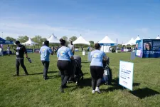 Thumbnail for Volunteers in blue shirts push strollers on grass at an outdoor event with white tents on a sunny day.