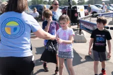 Thumbnail for Children receiving giveaway bags from Telhio Credit Union staff during an outdoor event on a sunny day.