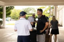 Thumbnail for Two men in casual golf attire talking under a covered outdoor area near a table with people and trees in background
