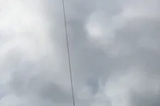 Thumbnail for Helicopter flying in cloudy sky with a long cable hanging down vertically from it.