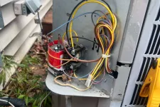 Thumbnail for Open outdoor HVAC unit showing wires, a red capacitor, and a yellow diagnostic tool attached during maintenance.
