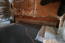 Thumbnail for Exposed crawl space with insulation, piping, and a dark floor panel beneath a wooden subfloor with shoes visible.