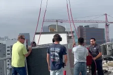 Thumbnail for Four HVAC technicians guide a large unit on a rooftop using a crane under a cloudy sky in an urban area.