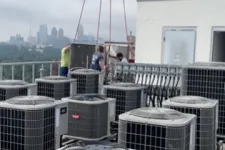 Thumbnail for Technicians installing HVAC units on a rooftop with city skyline and cloudy sky in the background.