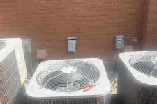 Thumbnail for Three outdoor air conditioning units against a red brick wall with a pair of red pliers on the center unit's top grille.