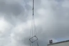 Thumbnail for Two workers on rooftop guiding a large metal frame suspended by crane cables in cloudy weather.