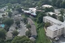 Thumbnail for Helicopter flying low above residential neighborhood with buildings and greenery under cloudy sky.